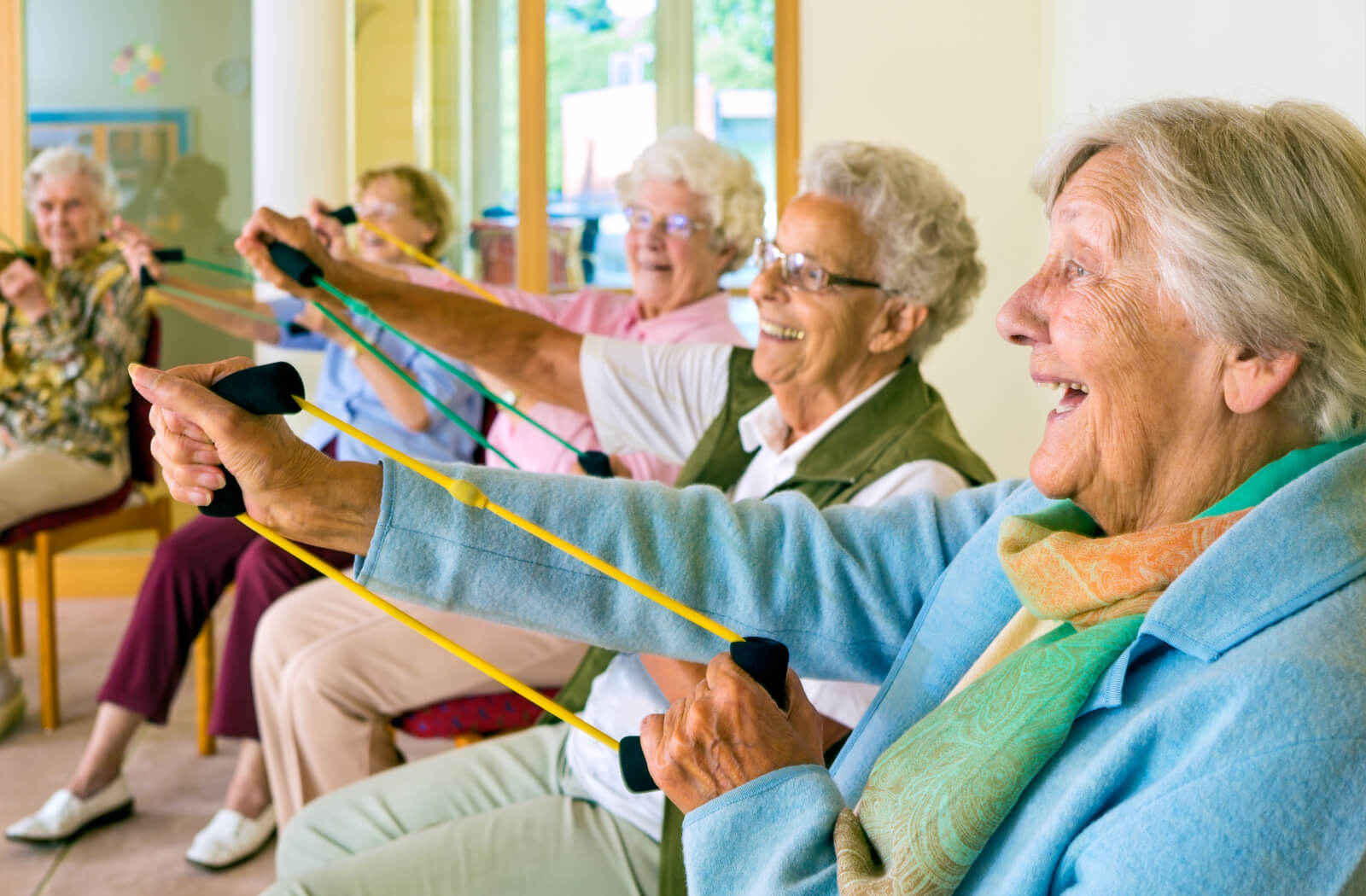A group of older adults laugh while stretching colorful resistance bands during an exercise class