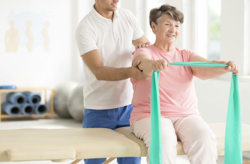 An older adult participates in stroke rehabilitation by stretching a resistance band with support from a physical therapist.
