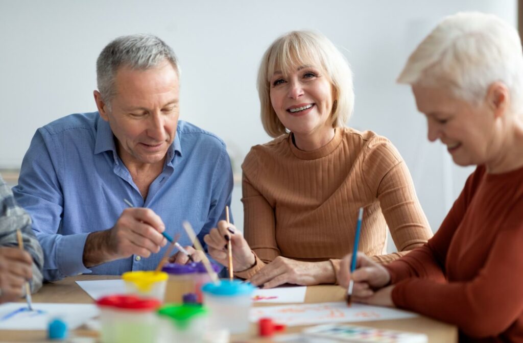Three older adults participating in a group painting activity at a memory care community, smiling and engaged in creative expression.